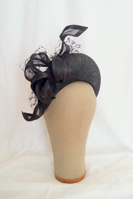 A black fascinator with a textured fabric base, large layered ribbon bows, and sheer netting, displayed on a beige mannequin head against a white fabric backdrop.