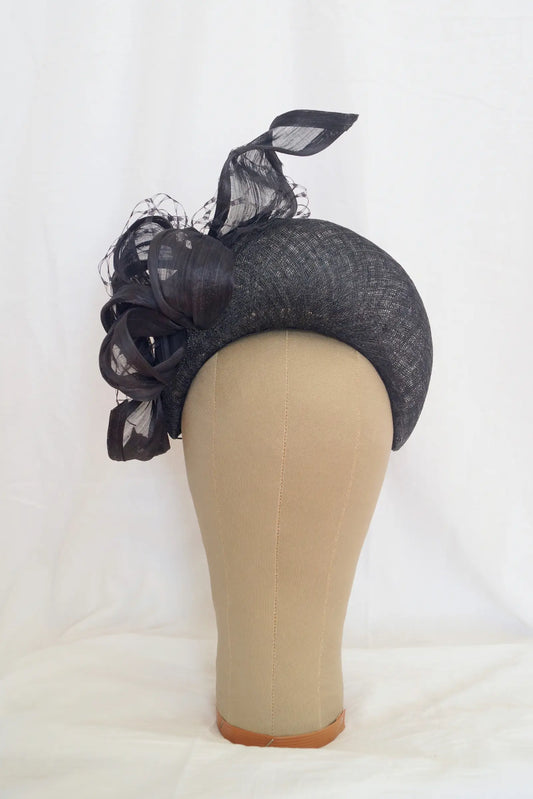 A black crescent shaped fascinator with matching black mesh and ribbon accents, displayed on a beige mannequin head against a white fabric backdrop.