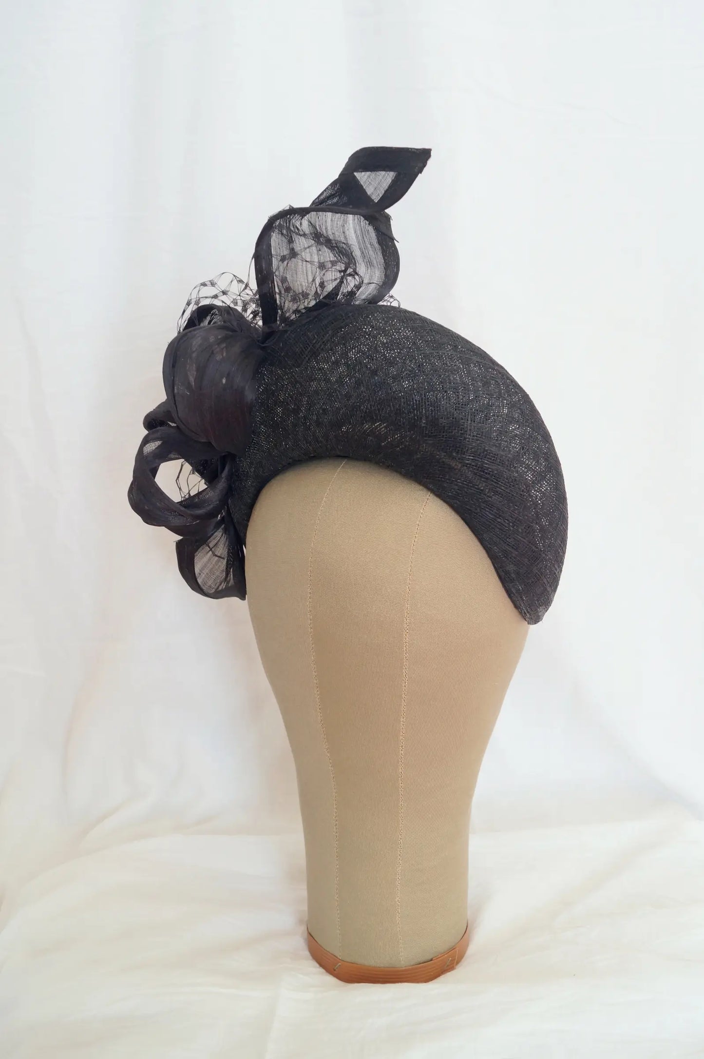 A black fascinator with sheer mesh and ribbons, displayed on a beige mannequin head against a white fabric backdrop.