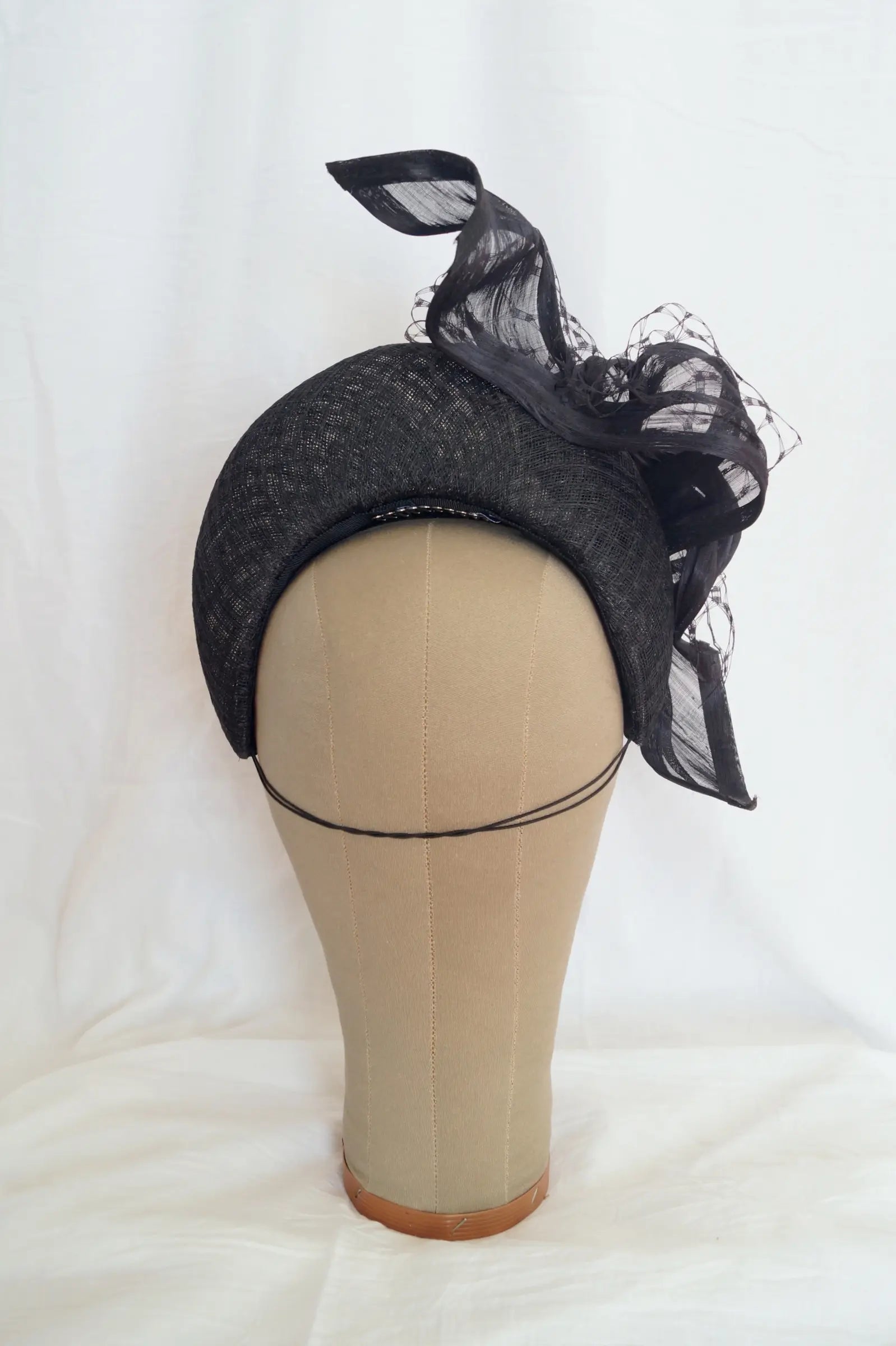 A black crescent shaped fascinator with a textured fabric base and sheer black mesh ribbon accents, displayed on a beige mannequin head against a white fabric backdrop.