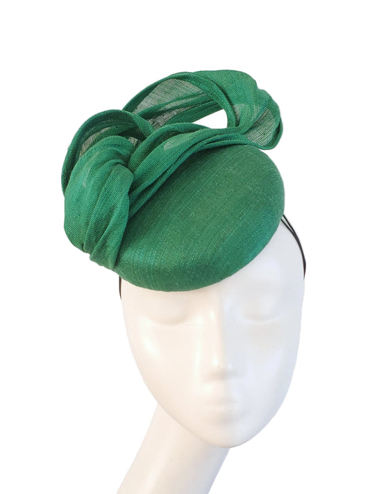 A green fascinator hat with a large sculptural bow and sheer mesh detailing, displayed on a white mannequin head against a white background.
