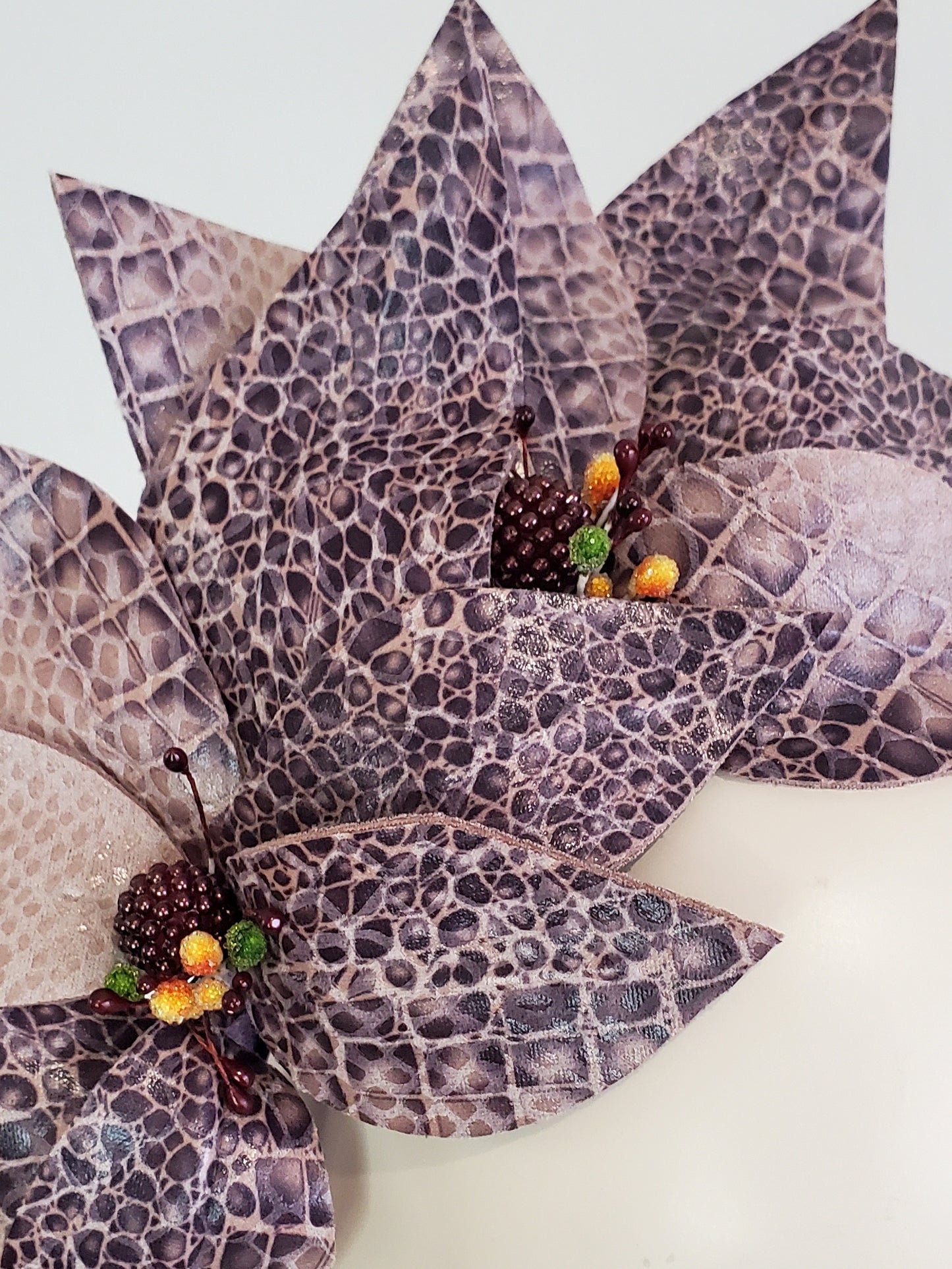 A close-up of a decorative floral arrangement featuring layered, petal-like pieces with a purple and gray reptile or snakeskin pattern, accented with small faux berries and green leaves on a plain light background.